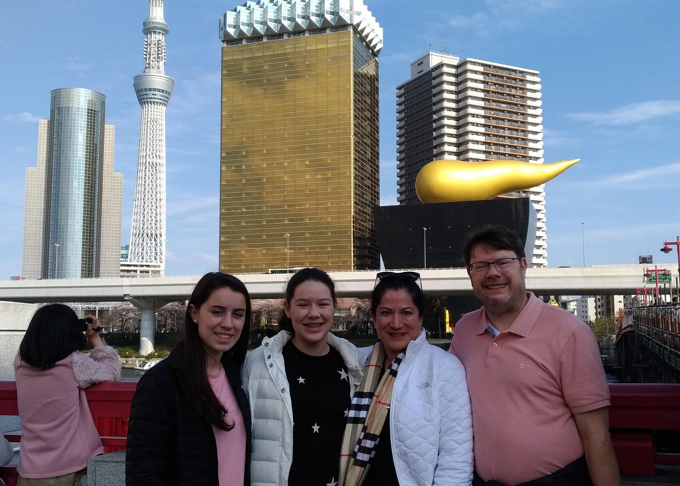 Travelers on a red bridge in Asakusa, with Tokyo Skytree and the golden Asahi flame behind them